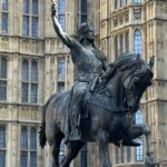 In front of an ornate Gothic-style building with many windows and decorative stonework stands a bronze equestrian statue. The man on the rearing horse raises his sword triumphantly, embodying a tale about movement and victory.
