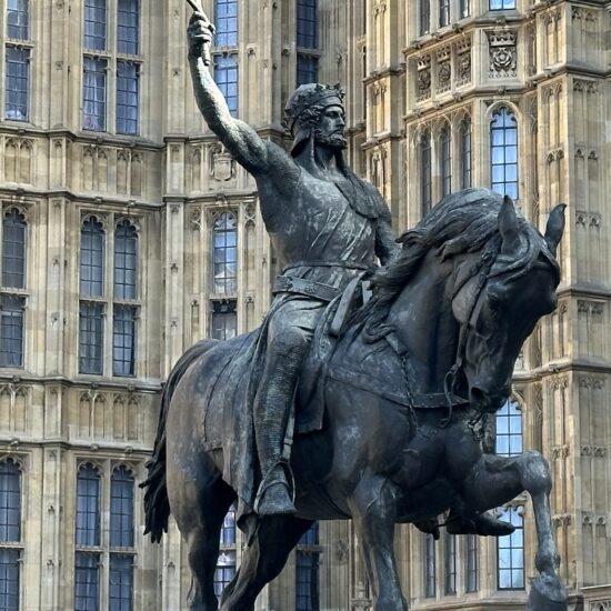 In front of an ornate Gothic-style building with many windows and decorative stonework stands a bronze equestrian statue. The man on the rearing horse raises his sword triumphantly, embodying a tale about movement and victory.
