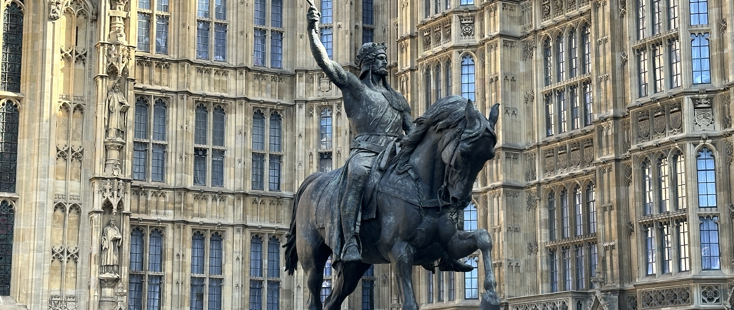 In front of an ornate Gothic-style building with many windows and decorative stonework stands a bronze equestrian statue. The man on the rearing horse raises his sword triumphantly, embodying a tale about movement and victory.