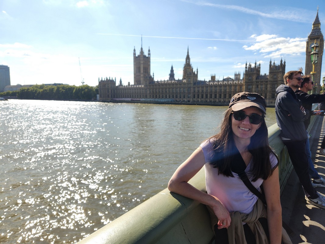 A woman wearing sunglasses and a cap smiles while standing on a bridge overlooking the River Thames. The Houses of Parliament and Big Ben are visible in the background under a sunny sky—a perfect moment on her London Christian Tour.