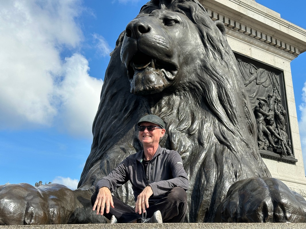 A person wearing sunglasses and a flat cap sits smiling at the base of a large lion statue, part of a monument with intricate carvings. Against the clear blue sky with clouds, this scene captures the charm of a London Christian tour.