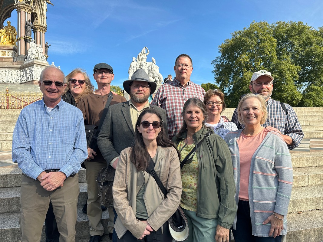 A group of ten people stands on a set of outdoor steps, smiling at the camera. They are on a London Christian Tour, posed in front of an ornate monument and surrounded by greenery under a clear, blue sky.