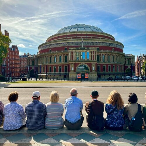 Eight people sit on a ledge, enjoying a London Christian Tour under the sunny sky. They face a large, round building with a domed roof, painted in red and brown with intricate details. Trees and other charming buildings dot the lively background.