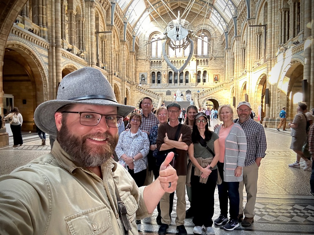 A group of people stands inside a grand, ornate museum with high arches, part of the London Christian Tour. A fossil skeleton is suspended from the ceiling, and in the foreground, someone takes a selfie and gives a thumbs-up.