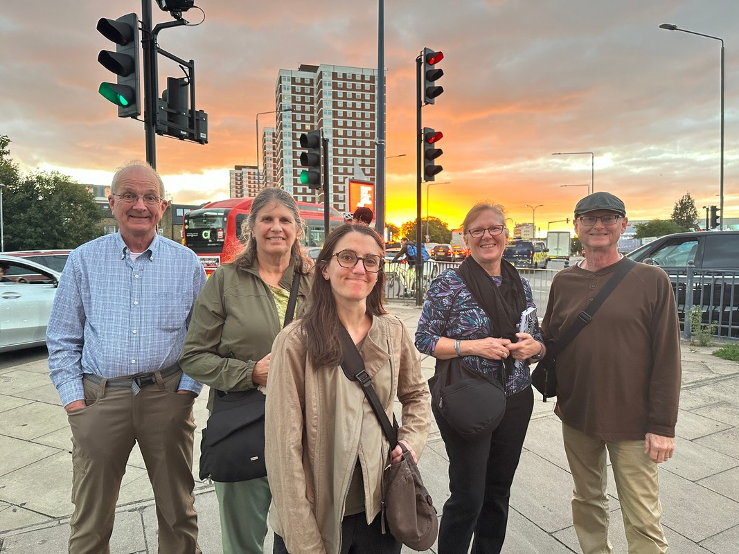 Five people stand at a bustling London street corner, the city buildings glowing under the sunset. Smiling and dressed casually, they share stories of their days unforgettable Christian tour. Nearby, traffic lights flicker and a red double-decker bus adds to the vibrant urban scene.