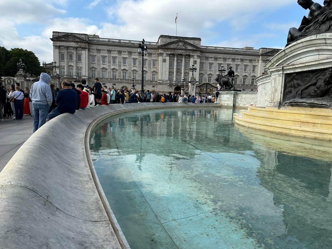 A crowd of people lines up outside Buckingham Palace, with the palace in the background. On a London Christian Tour, visitors often pause by the curved stone ledge around a reflective pool of water, gazing up at the cloudy sky above as they soak in the historical ambiance.