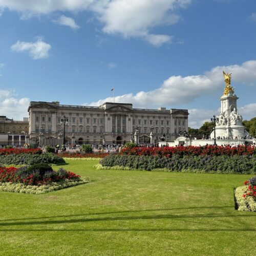 The image captures Buckingham Palace on a sunny day in London, with a clear blue sky above. In the foreground, lush green lawns are adorned with colorful flower beds, creating a serene backdrop for those on a London Christian Tour. The iconic Victoria Memorial stands prominently on the right side.