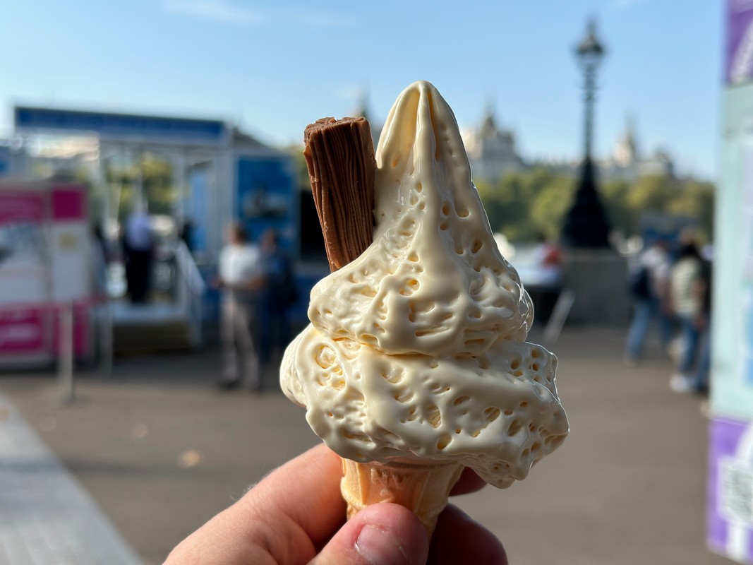 A person holds a creamy soft-serve ice cream cone with a chocolate flake on top, enjoying a sweet moment during a London Christian tour. The background shows a blurred outdoor scene with people and historic buildings under the clear blue sky.