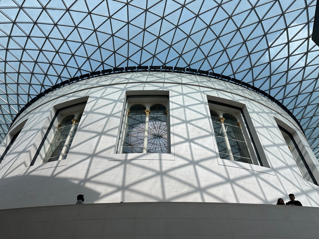 A round white building with large windows under a glass dome roof in London. The shadows of the geometric grid pattern on the roof are cast on the walls, creating a mesmerizing effect. As part of the Christian tour, two people are visible at the bottom right corner, marveling at its architecture.