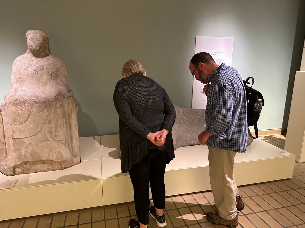 Two people examine ancient stone sculptures in a museum, part of the London Christian Tour. One figure is seated while the other reclines. The visitors lean in, showing interest and discussion, with their backs to the camera. The room features neutral-colored walls and tiled flooring.