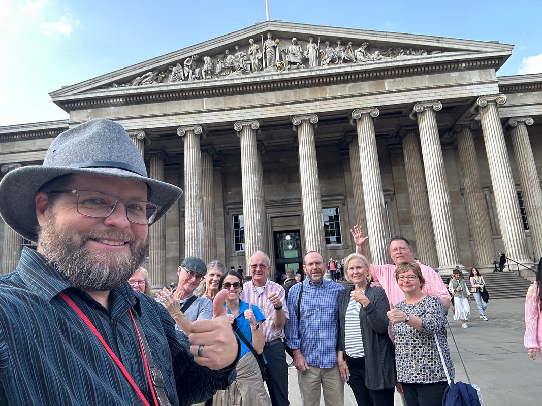A group of people posing in front of the British Museum building with tall columns and ornate sculptures on the pediment, capturing memories from their London Christian Tour. A person in a hat takes a selfie with others smiling and waving in the background.