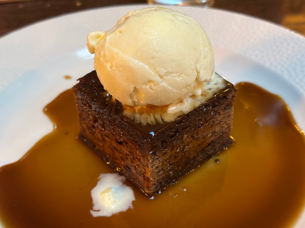 A close-up of a sticky toffee pudding served on a plate, topped with a scoop of vanilla ice cream. Reminiscent of London’s sweet culinary delights, the dessert is surrounded by a pool of rich caramel sauce.