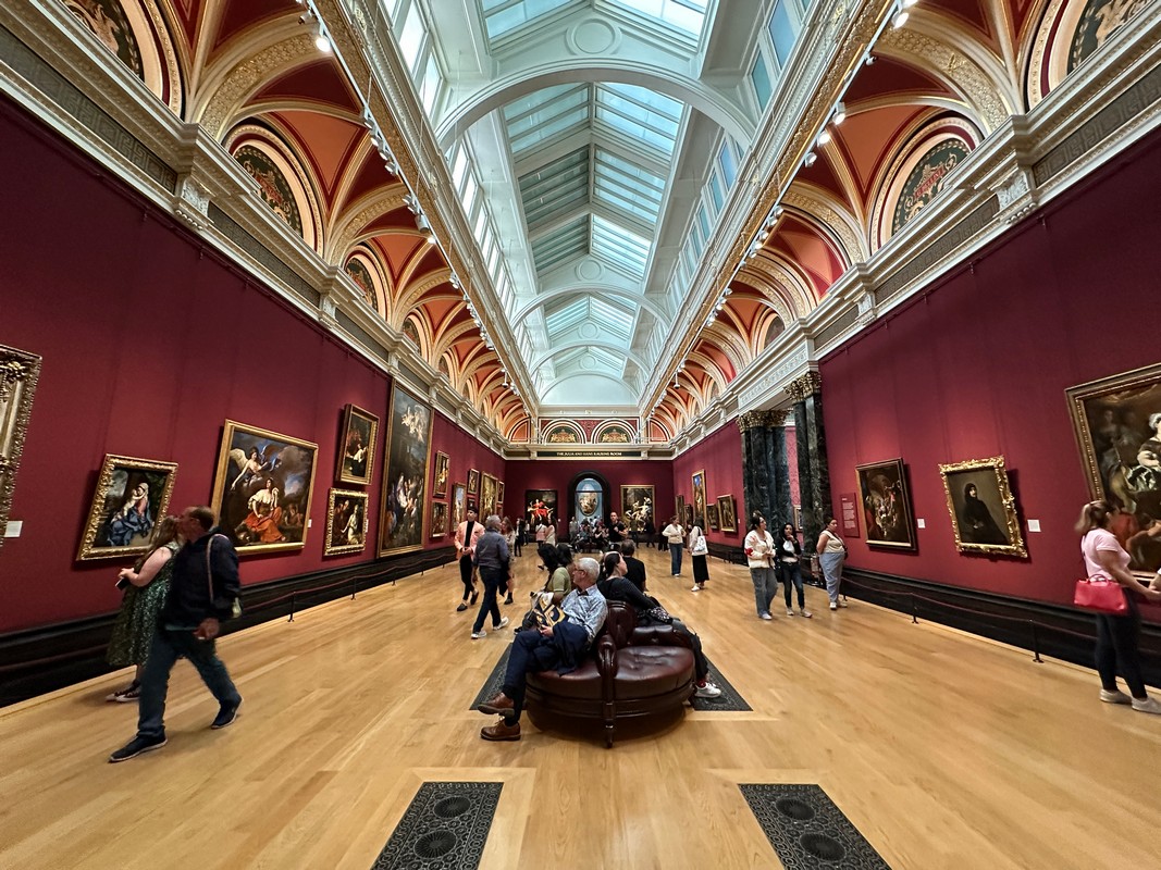 A spacious art gallery with high arched ceilings and a skylight. Visitors on the London Christian Tour admire portraits and classical paintings on deep red walls. Central seating area with people resting, while others walk and observe the artwork.