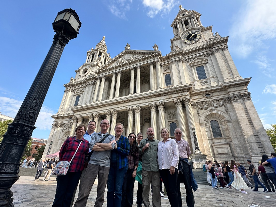 A group of eight people stands in front of a large, historic cathedral, featuring tall columns and two clock towers during their London Christian Tour. A lamp post is visible on the left, and a wedding party can be seen in the background. Its a sunny day with a few clouds.