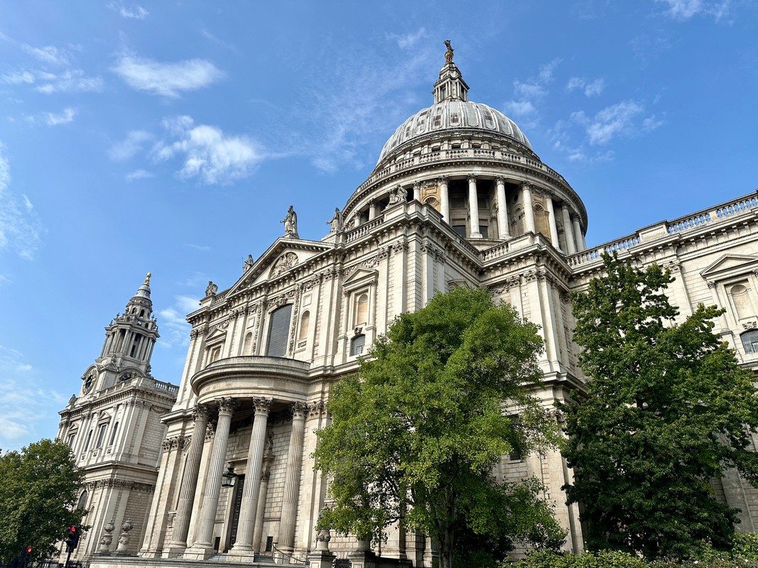 St. Pauls Cathedral in London majestically rises, its grand dome and classical architecture highlighted on a blue sky with scattered clouds. Green trees partially obscure the view of this historic landmark, a must-see on any London Christian Tour exploring the citys rich religious heritage.