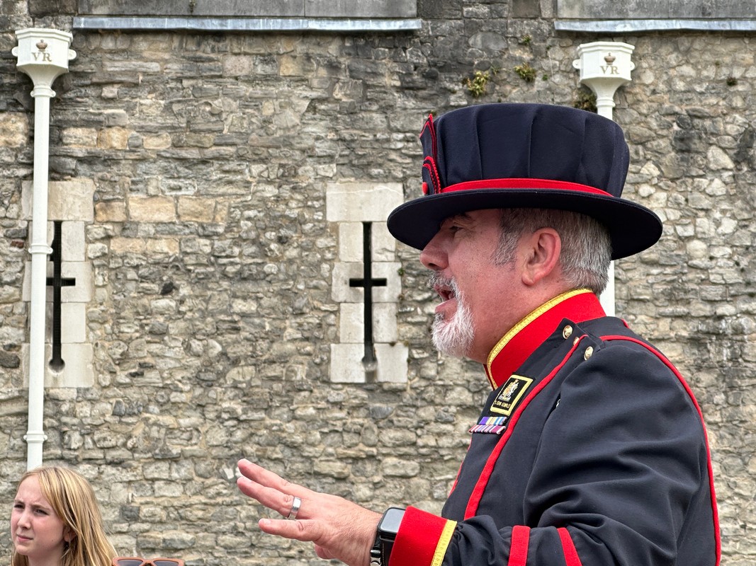 A Beefeater in traditional uniform, including a black hat with red trim, speaks outdoors in front of a stone wall during a London Christian Tour. A few unidentified people are partially visible in the foreground, adding to the historical ambience.