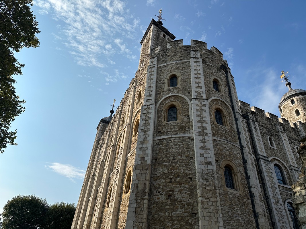 A historic stone tower, once part of a medieval fortress, rises proudly against a blue sky with scattered clouds. The structure, featured on the London Christian Tour, boasts arched windows and a crenellated roof. Lush trees accentuate the foreground.