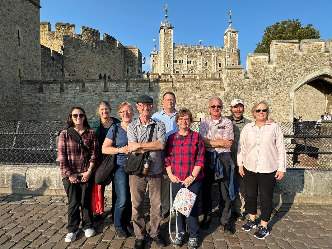 A group of nine people smiling and posing in front of the historic Tower of London, enjoying a sunny day with clear blue skies. The medieval architecture forms a stunning backdrop for this moment on their London Christian Tour.