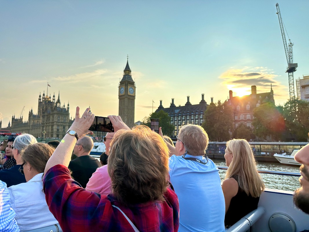 People are on a London Christian Tour boat ride during sunset, capturing photos of the Houses of Parliament and Big Ben. The sun casts a warm glow on the scene, and a crane is visible in the background.