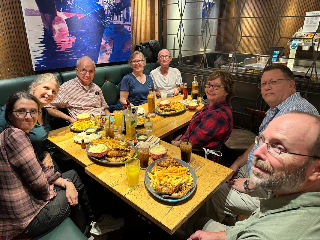 A group of eight people sits around a wooden table at a London Nando's in Earl's Court, sharing a meal. The table is filled with plates of food, drinks, and condiments. The atmosphere is warm, and the group appears to be enjoying their time together on their Christian tour..