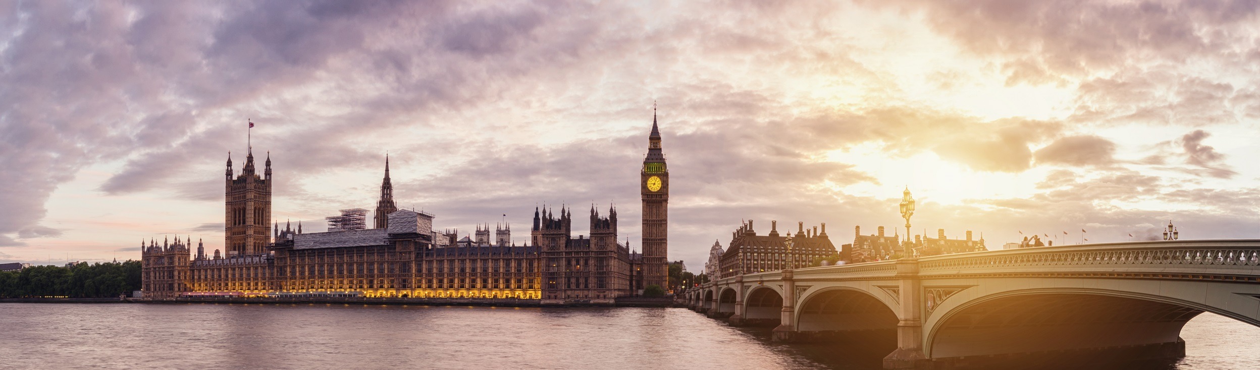 Panoramic view of the Houses of Parliament and Big Ben in London at sunset, with the River Thames in the foreground and Westminster Bridge to the right. The sky is partly cloudy with a warm glow from the setting sun.