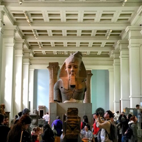 Visitors on their tour itinerary explore a museum hall with a large ancient Egyptian statue, likely of a pharaoh, prominently displayed in the center. The rooms high ceilings and architectural detailing captivate tourists as they observe and take photos.