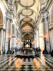 St-Pauls-Cathedral-London The grand cathedral interior, often featured in tour itineraries, boasts high arched ceilings, ornate chandeliers, and marble columns. Visitors wander across the black and white checkered floor, marveling at the majestic architecture.