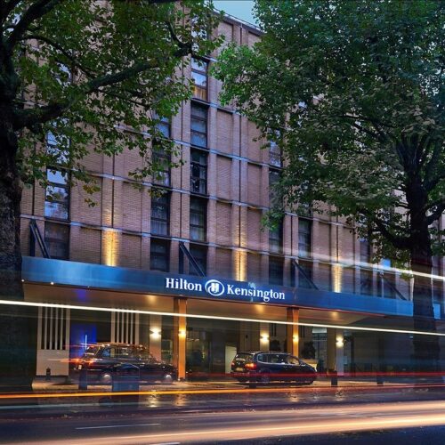 Street view of Hilton London Kensington at dusk, a perfect start for any tour itinerary. The hotel features a modern facade with large trees in front. Blurred lights from passing cars create streaks, and a few taxis are parked nearby.