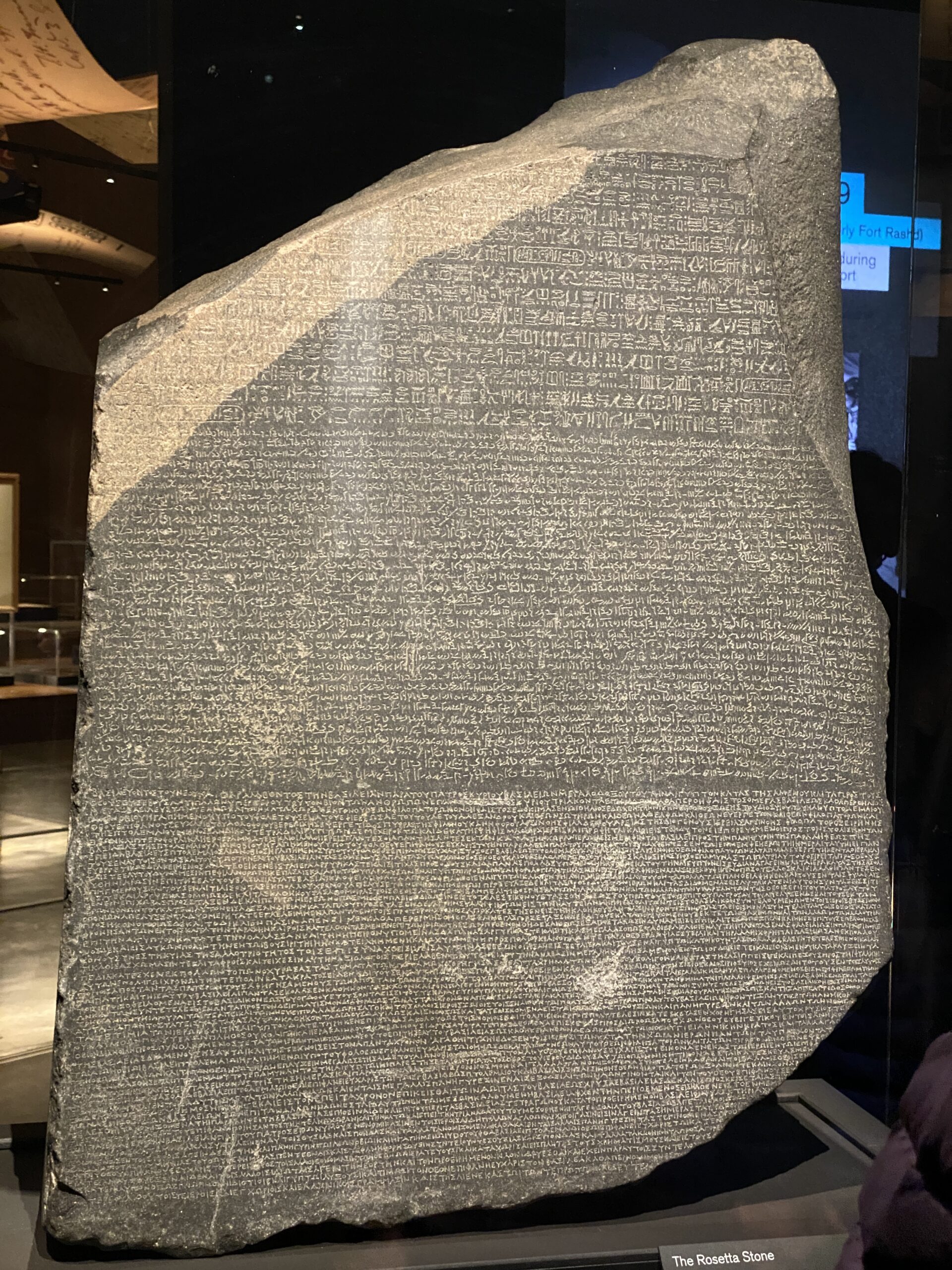 The Rosetta Stone, a large dark stone slab with inscriptions in three scripts—hieroglyphic, Demotic, and Ancient Greek—is displayed upright under glass in a renowned Christian London museum exhibit.