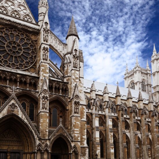 Gothic-style exterior of Westminster Abbey in London, featuring intricate stonework reminiscent of the detail seen on the Rosetta Stone, arched doorways, a large rose window, pointed spires, and tall towers under a partly cloudy blue sky.