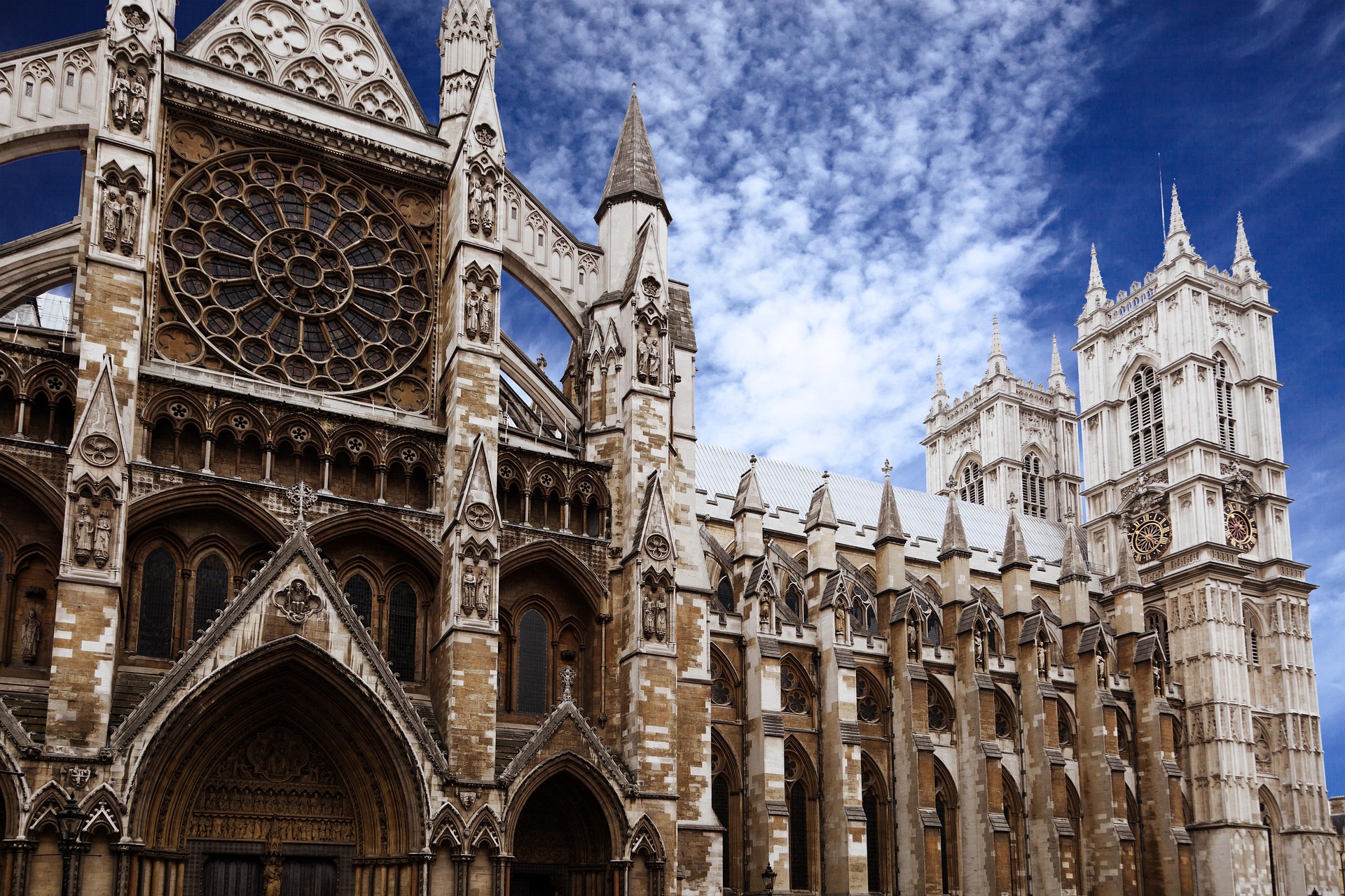 Gothic-style exterior of Westminster Abbey in London, featuring intricate stonework reminiscent of the detail seen on the Rosetta Stone, arched doorways, a large rose window, pointed spires, and tall towers under a partly cloudy blue sky.
