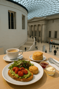 A table set with avocado toast, salad, scones, jam, clotted cream, and a cup of tea overlooks the spacious, modern interior of a museum with a glass roof and visitors below.