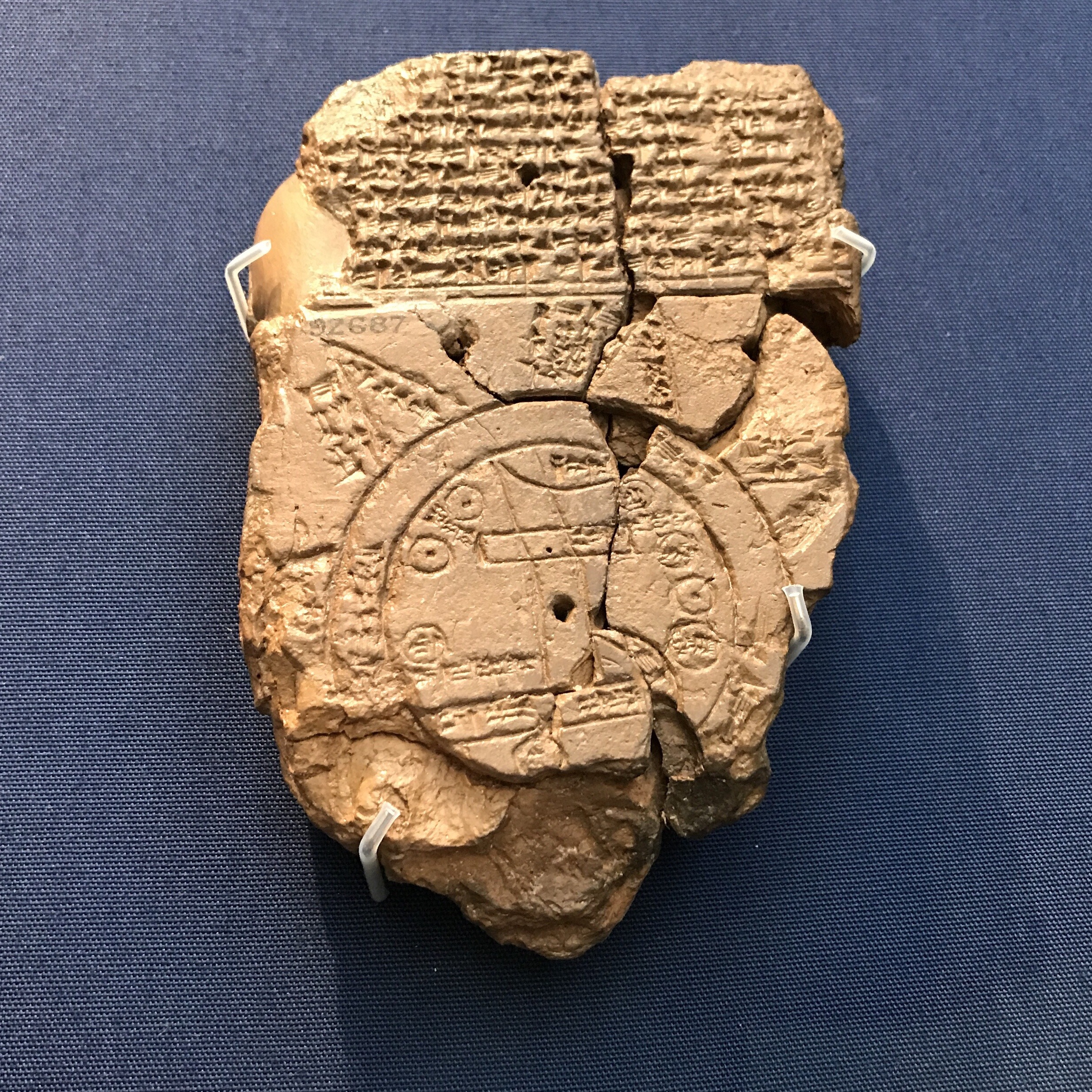 Babylonian Map of the World in the British Museum - Nate Loper. A broken ancient clay tablet inscribed with cuneiform text and a circular map-like diagram, displayed on a blue background with small supports holding the fragments together.