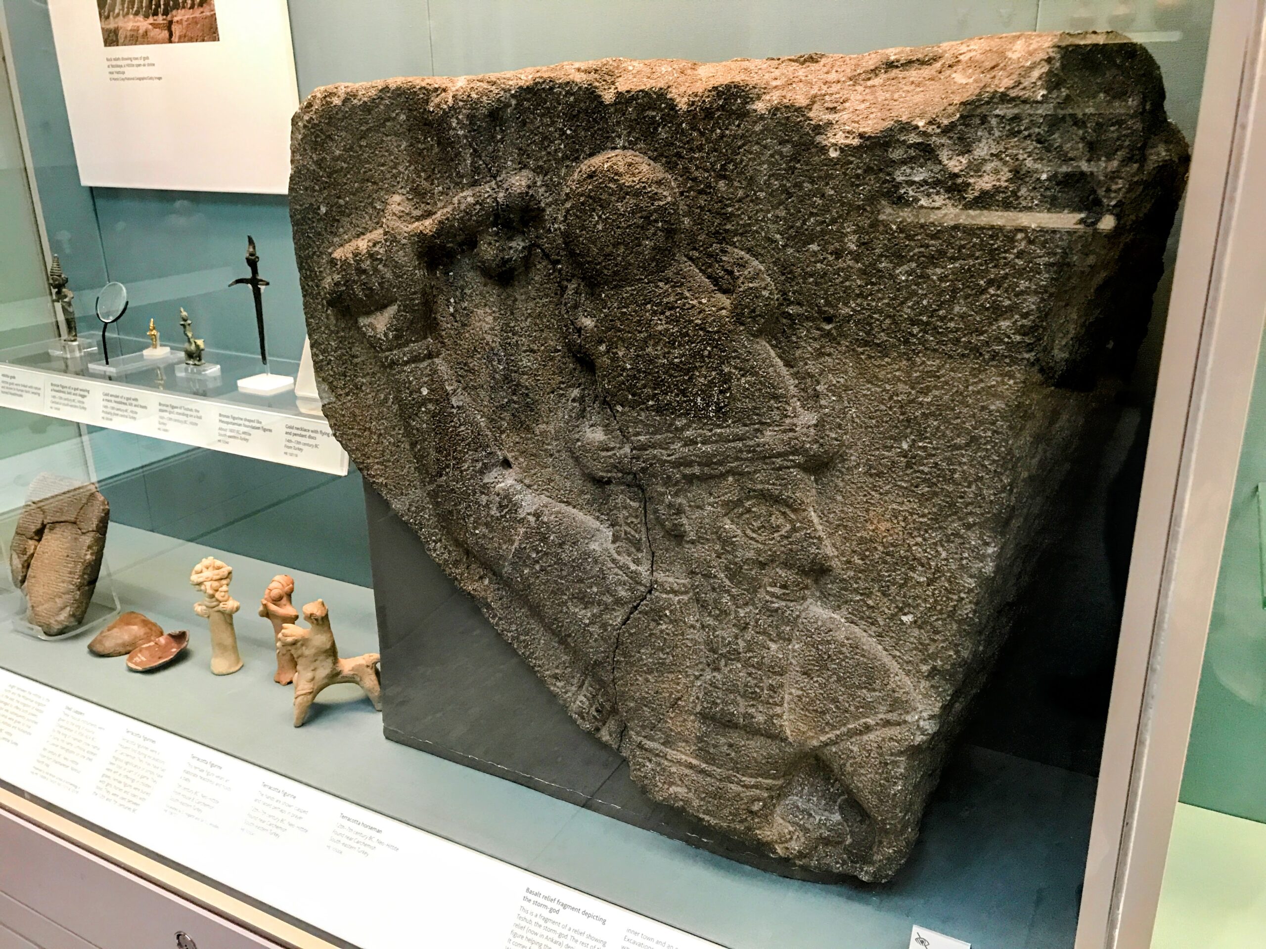Hittite Culture in the British Museum, Nate Loper. A stone relief depicting a figure wearing a headdress and holding a weapon, displayed in a museum case with other ancient artifacts and descriptive labels. London Christian Tours