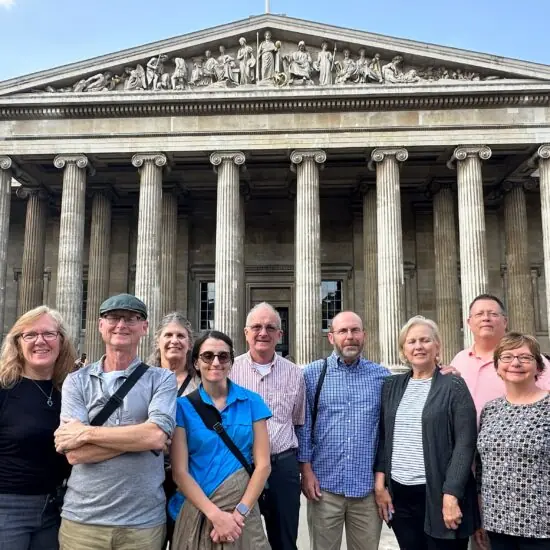 A group of nine adults stands smiling in front of the British Museum’s grand entrance with tall columns and classical pediment on a sunny day.