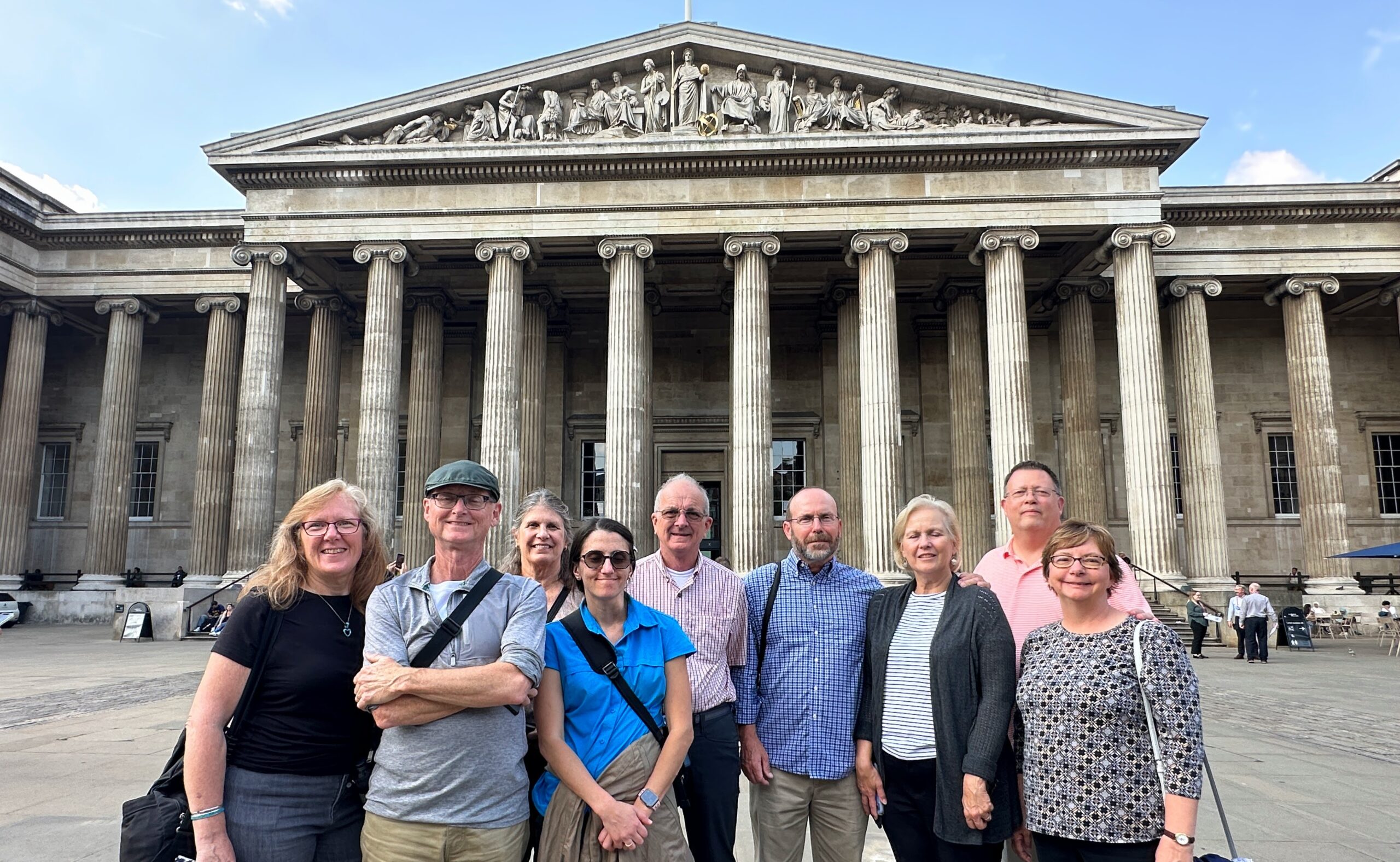 A group of nine adults stands smiling in front of the British Museum’s grand entrance with tall columns and classical pediment on a sunny day.