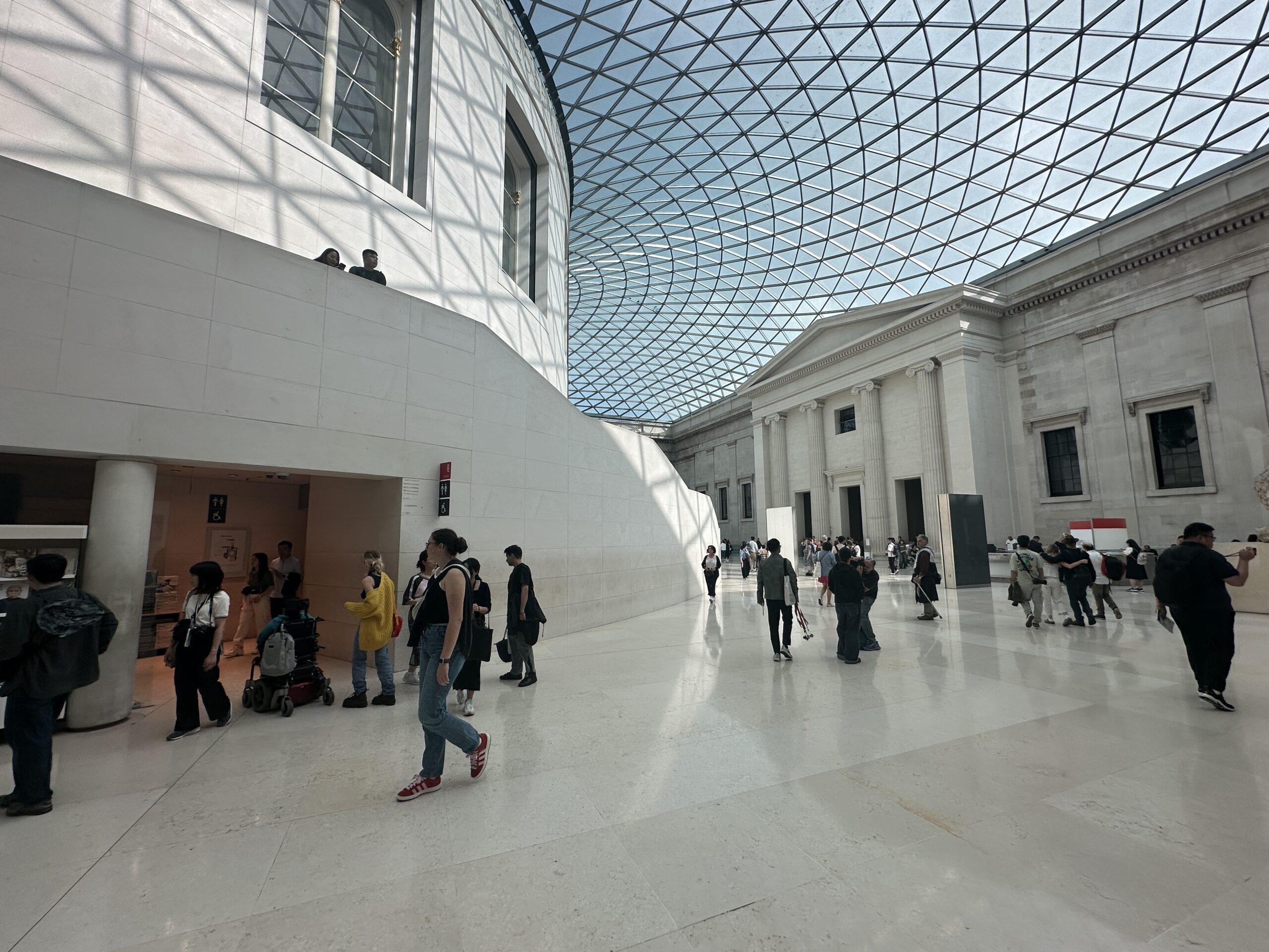 British Museum Courtyard. People walk and gather in a spacious, bright atrium with a glass geometric ceiling and white stone walls, likely inside a museum or gallery. Sunlight streams through the roof, casting shadows on the floor.