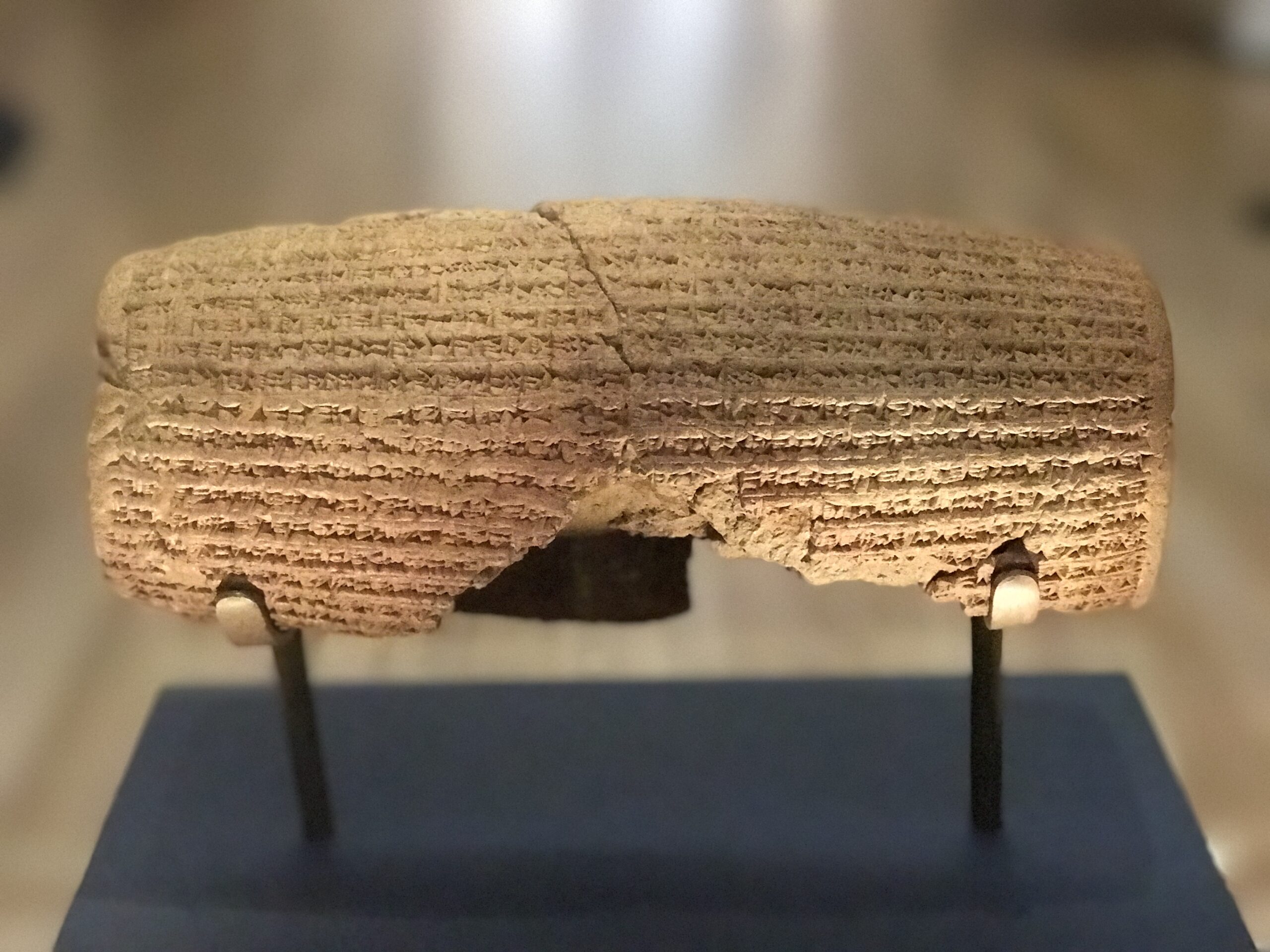 The Cyrus Cylinder in the British Museum by Nate Loper. A cylindrical clay artifact with ancient cuneiform script carved on its surface, displayed on a stand in a museum setting. London Christian Tours